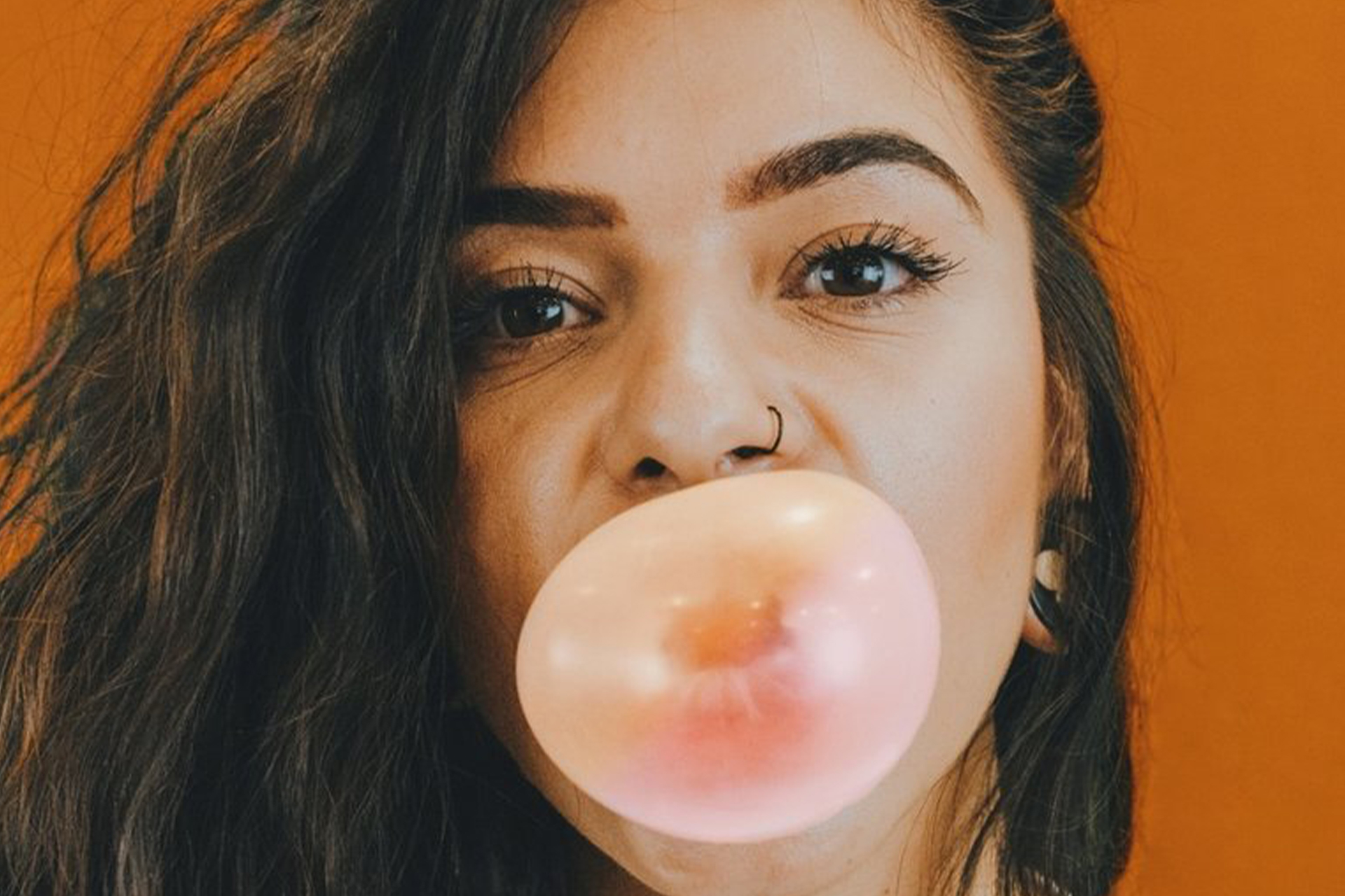 A young woman with a nose ring blowing a Bubble gum bubble.