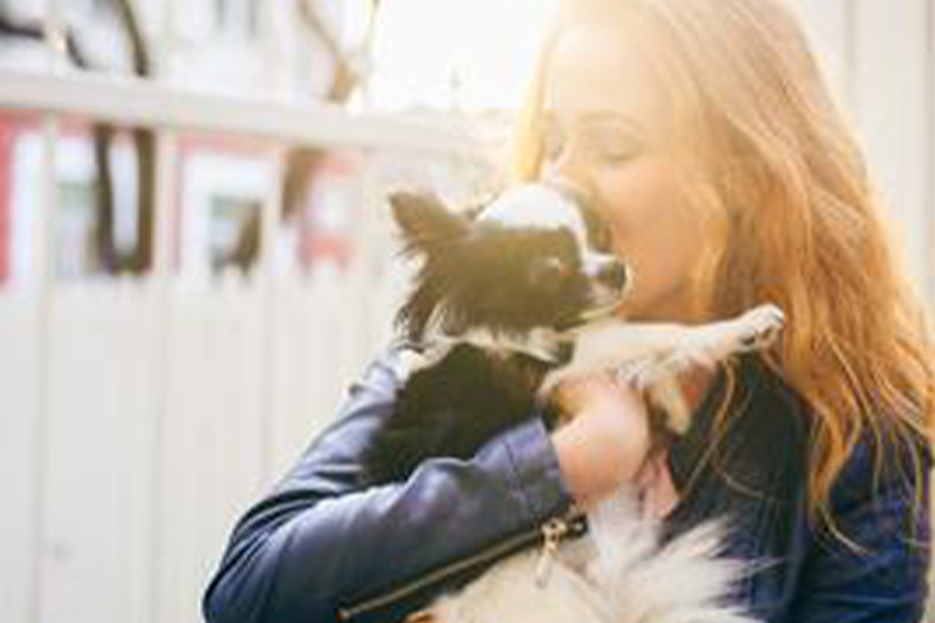 A young lady wearing a blue jacket holding and kissing a small black and white dog.