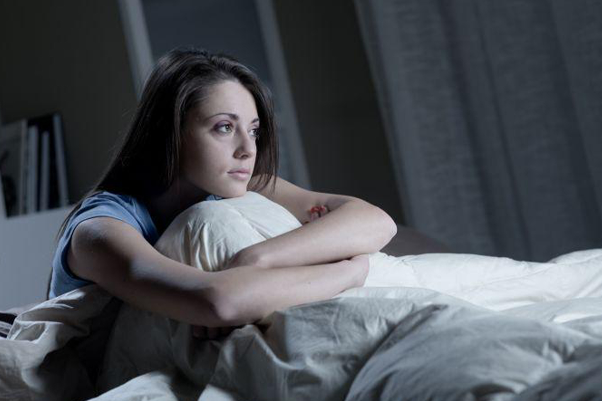 A young woman sitting in bed looking into the distance looking depressed, cradling her legs.