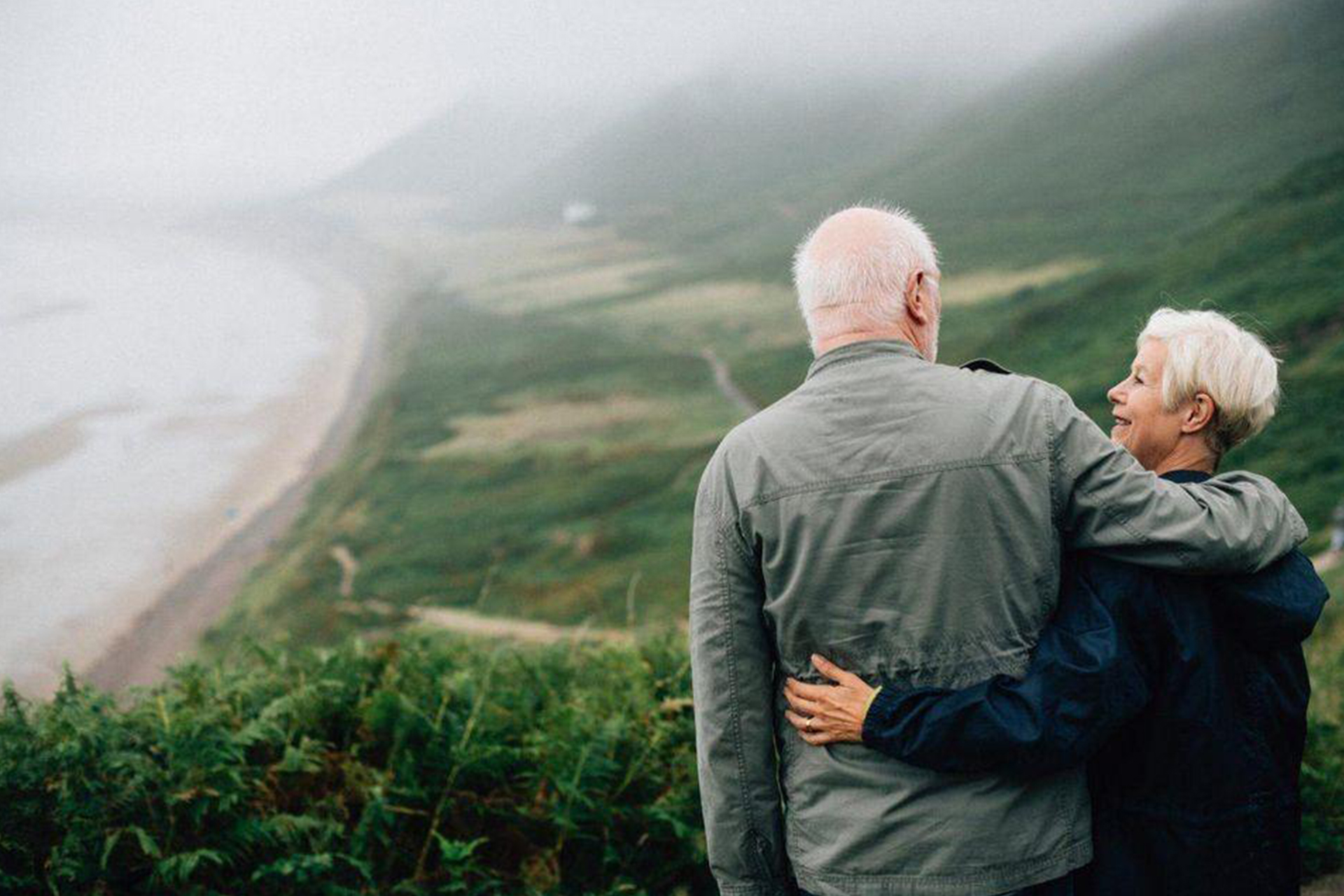 The back of an older couple embracing while looking over a mountain view.