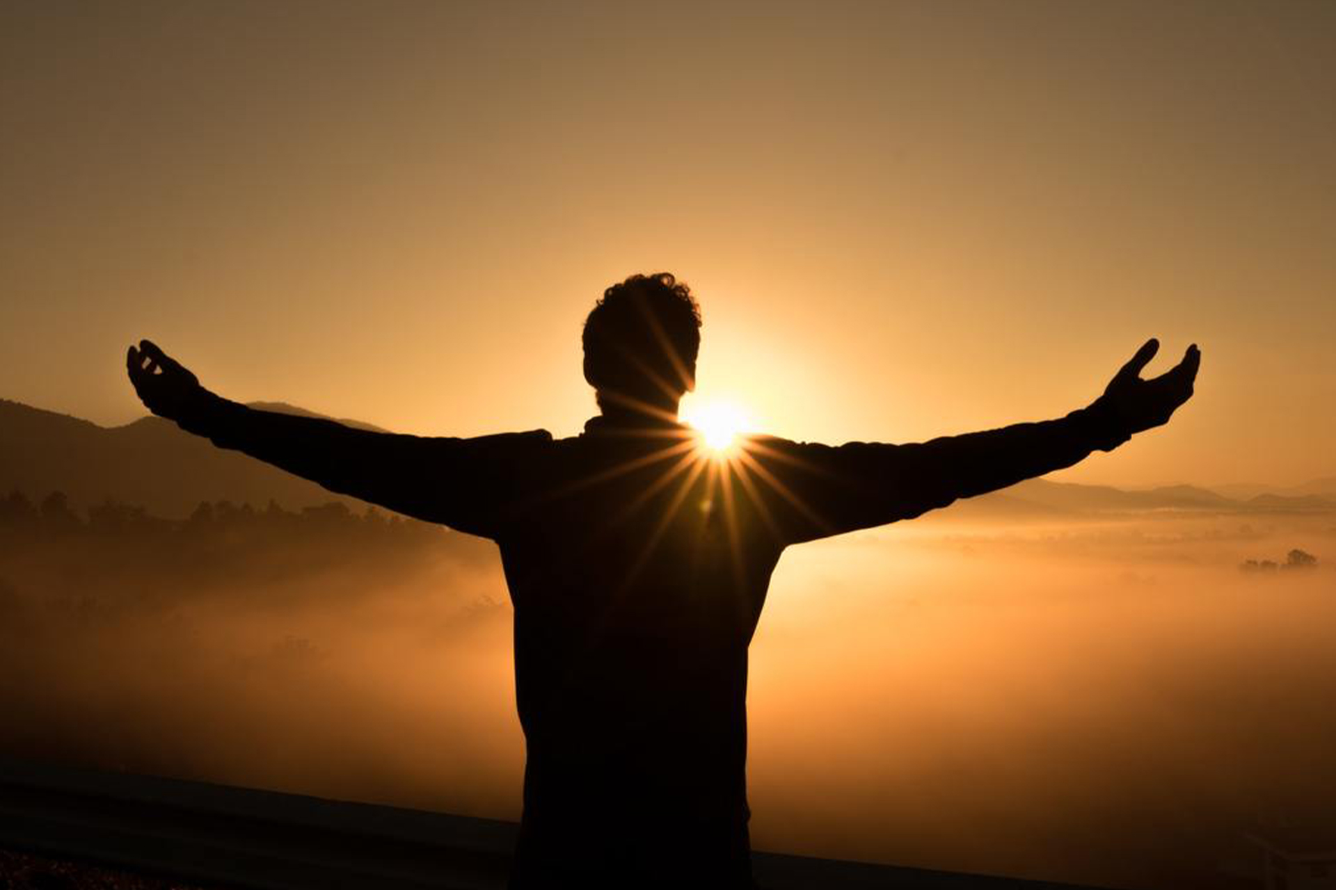 A man with outstretched arms looking over a landscape, with the setting sun directly in front of him.