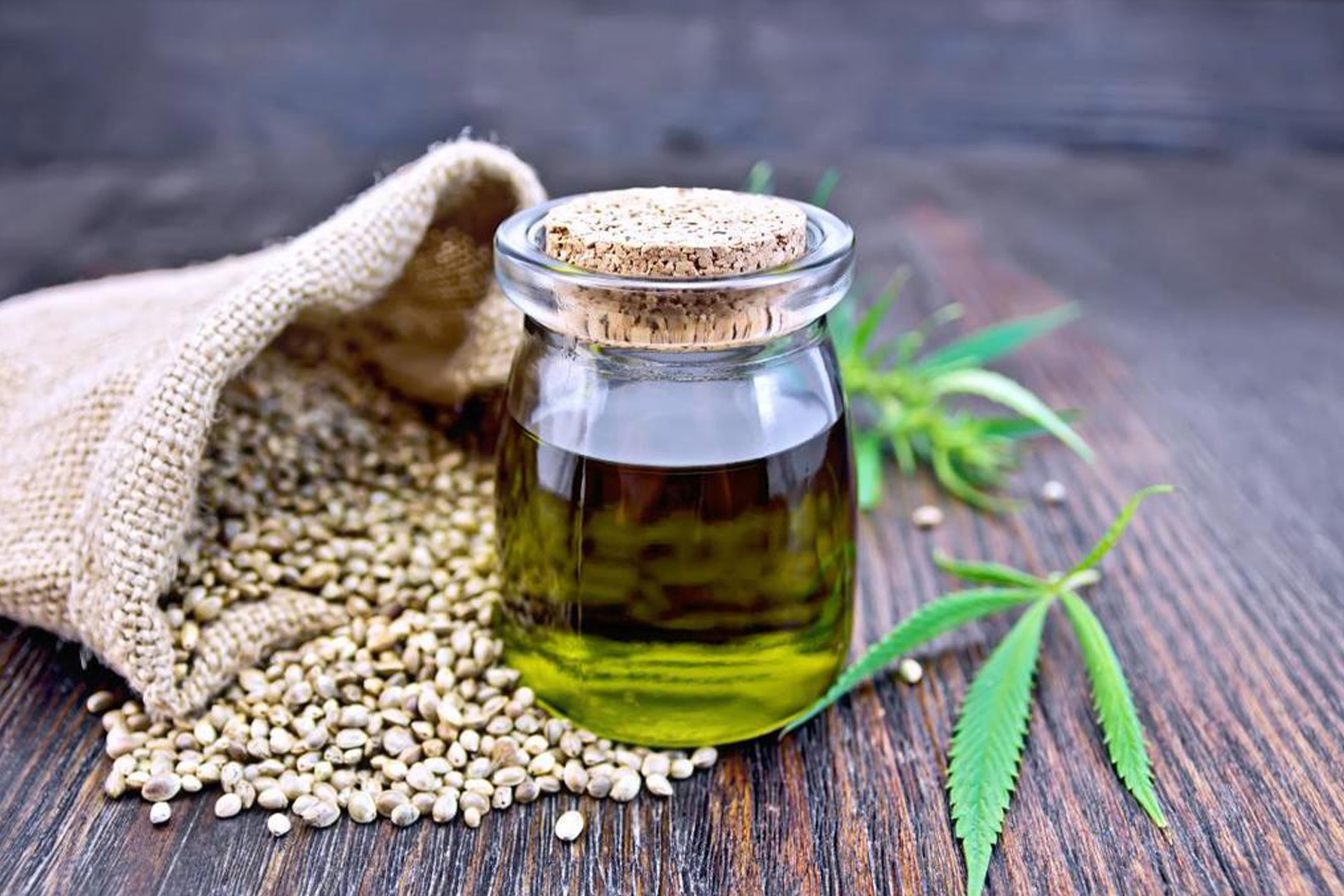 A glass jar with a cork lid containing CBD oil is placed on a wooden surface among a bag of Hemp Seeds that have spilled out, and two green Marijuana leaves to the side.